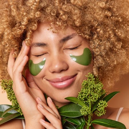 Photo of pleased young womann with curly bushy hair keeps hand on face smiles happily has eyes closed holds green plant applies beauty patches under eyes uses natural cosmetic. Skin care concept